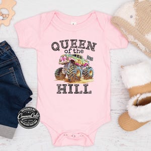 May include: A pink baby bodysuit featuring the text "QUEEN of the HILL" and a monster truck graphic. The monster truck is green, pink, and blue with large tyres. The bodysuit has snap closures.