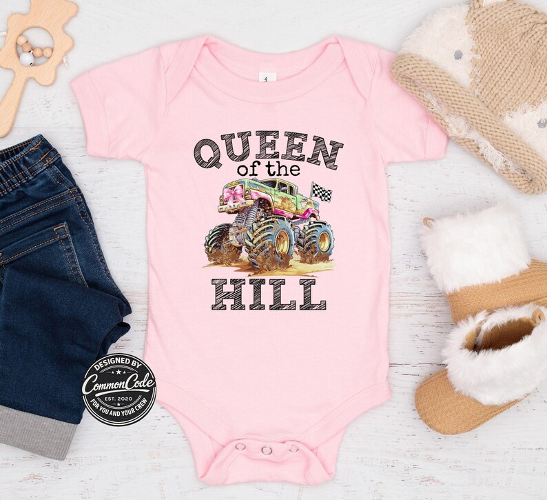 May include: A pink baby bodysuit featuring the text "QUEEN of the HILL" and a monster truck graphic. The monster truck is green, pink, and blue with large tyres. The bodysuit has snap closures.