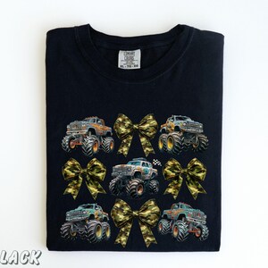 May include: Black t-shirt featuring a design of monster trucks and camouflage bows. The trucks are in various colors, with large tires. The bows are in a camouflage pattern. The text "BLACK" is at the bottom left.