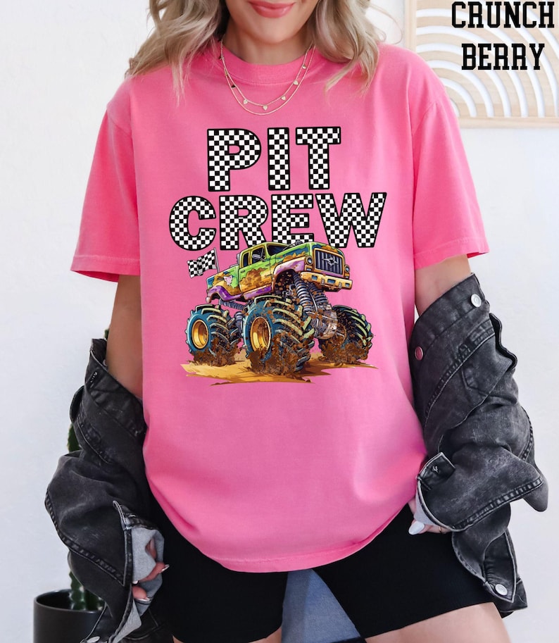 Lady is wearing a pink comfort colors t-shirt with a black and white checkered "PIT CREW" graphic and a green and purple cartoon monster truck with huge tires.
