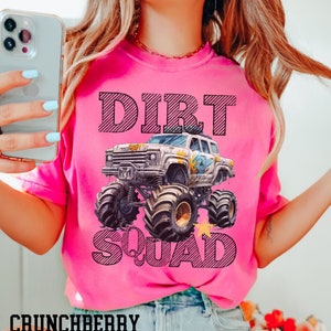Puede incluir: Una camiseta rosa brillante con las palabras "DIRT SQUAD" y un gr&aacute;fico de un monster truck. El texto y el dise&ntilde;o del cami&oacute;n son en negro. La palabra "CRUNCHBERRY" est&aacute; impresa en la parte inferior de la camiseta.