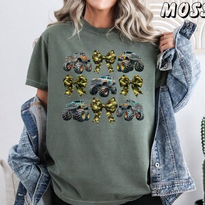 May include: Olive green t-shirt featuring a design of monster trucks and camouflage bows. The trucks are in various colors, and the bows add a decorative touch. The word "MOSS" is printed in the upper right corner.