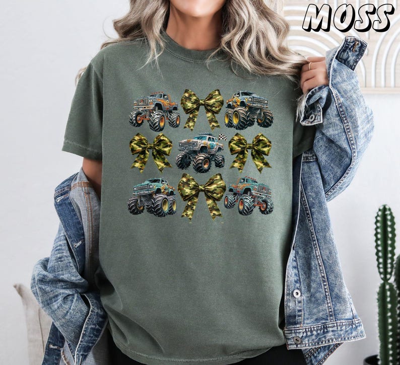 May include: Olive green t-shirt featuring a design of monster trucks and camouflage bows. The trucks are in various colors, and the bows add a decorative touch. The word "MOSS" is printed in the upper right corner.