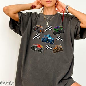 May include: Dark gray t-shirt featuring colorful monster truck graphics with checkered flags. The trucks are in various colors, including brown, green, red, blue, and orange. The shirt has a relaxed fit.
