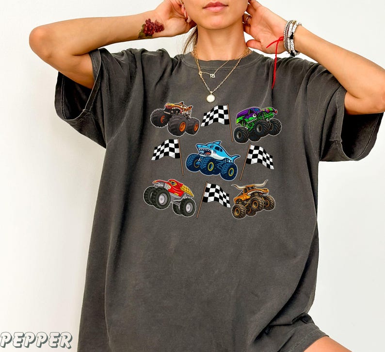 May include: Dark gray t-shirt featuring colorful monster truck graphics with checkered flags. The trucks are in various colors, including brown, green, red, blue, and orange. The shirt has a relaxed fit.