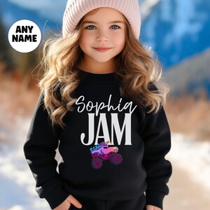 May include: Black sweatshirt with the name "Sophia" and the word "JAM" in white. A colorful unicorn monster truck graphic is below the word "JAM". The sweatshirt is worn by a person wearing a pink beanie.