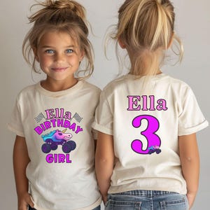 Pictured is a young girl wearing a beige natural colored t-shirt 2-sided design which says Ella Birthday Girl in pink and purple letters with a unicorn sparkle smash monster truck and 2 checkered flags. on the back there is custom name Ella a big number 3 and a small version of the truck graphic