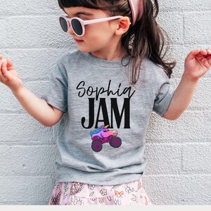 May include: A gray t-shirt with the text "Sophia JAM" in black script and block letters. Below the text is a colorful monster truck graphic. The shirt is worn by a young child wearing pink sunglasses and a pink headband.