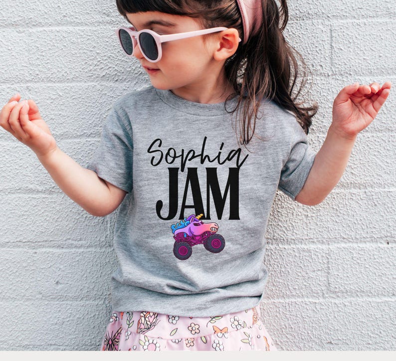 May include: A gray t-shirt with the text "Sophia JAM" in black script and block letters. Below the text is a colorful monster truck graphic. The shirt is worn by a young child wearing pink sunglasses and a pink headband.