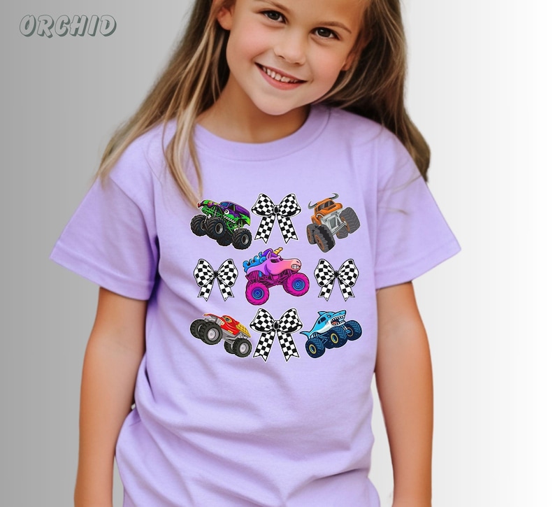 May include: A light purple t-shirt featuring colorful monster truck graphics and checkered bows. The trucks are in various colors, including green, orange, red, blue, and pink. The word "ORCHID" is in the upper left corner.