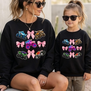 Op de afbeelding: Zwarte sweatshirts met kleurrijke monster truck afbeeldingen en roze strikaccenten. Een vrouw en een kind dragen de sweatshirts, wat wijst op een bijpassende set. De monster trucks zijn in diverse kleuren.