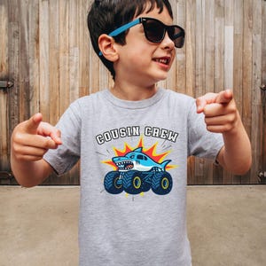 model is wearing a grey t-shirt that says "Cousin Crew" with a shark monster truck below. Any Custom Name, Family Matching Made to match. Adult, kid, toddler, youth and baby sizes.