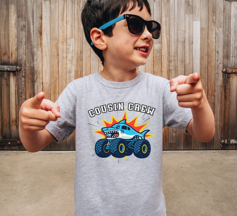 model is wearing a grey t-shirt that says "Cousin Crew" with a shark monster truck below. Any Custom Name, Family Matching Made to match. Adult, kid, toddler, youth and baby sizes.