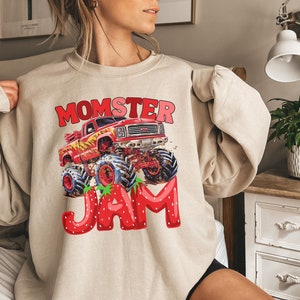 May include: Beige sweatshirt with a graphic featuring a red monster truck with flames, the word "MOMSTER" in red and pink, and the word "JAM" in strawberry-shaped letters. The design is centered on the front of the sweatshirt.