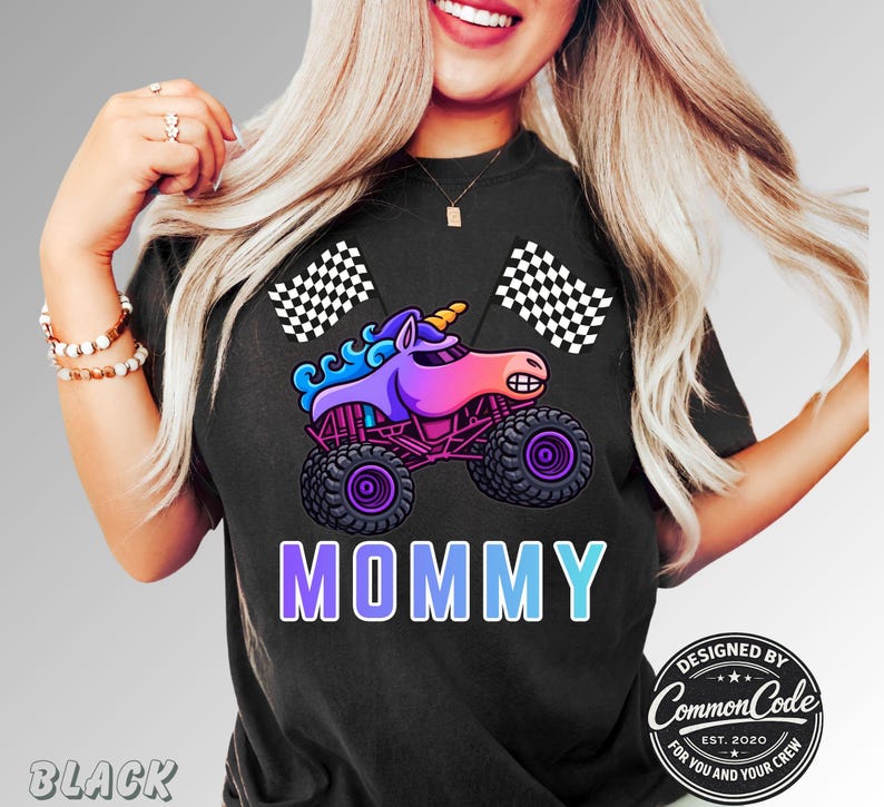 Mom is wearing a Black t-shirt featuring a colorful graphic of a unicorn sparkle monster smash truck with the word "MOMMY" in a blue gradient font. Two checkered flags are on either side of the truck. The design includes the text "DESIGNED BY COMMON CODE EST. 2020 FOR YOU AND YOUR CREW."