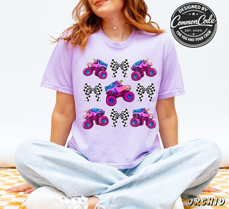 A woman is wearing a light purple lavender colored t-shirt featuring a pattern of monster trucks with unicorn heads and checkered bows. The shirt has the text "DESIGNED BY Common Code EST. 2020 FOR YOU AND YOUR CREW" in a circular design.