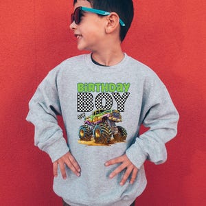 May include: A light gray sweatshirt with a colorful monster truck graphic. The text "BIRTHDAY BOY" is printed above the truck in green and black checkered font. The boy in the image is wearing sunglasses and smiling.