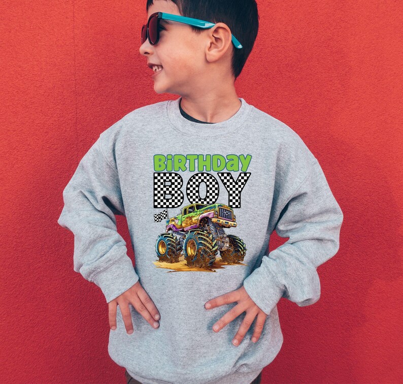 May include: A light gray sweatshirt with a colorful monster truck graphic. The text "BIRTHDAY BOY" is printed above the truck in green and black checkered font. The boy in the image is wearing sunglasses and smiling.