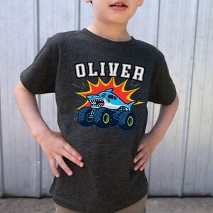 May include: Dark gray t-shirt featuring the name "OLIVER" in white block letters above a cartoon shark monster truck design. The shark is blue and white with sharp teeth, and the truck has large blue wheels. The design is set against a red and yellow burst.