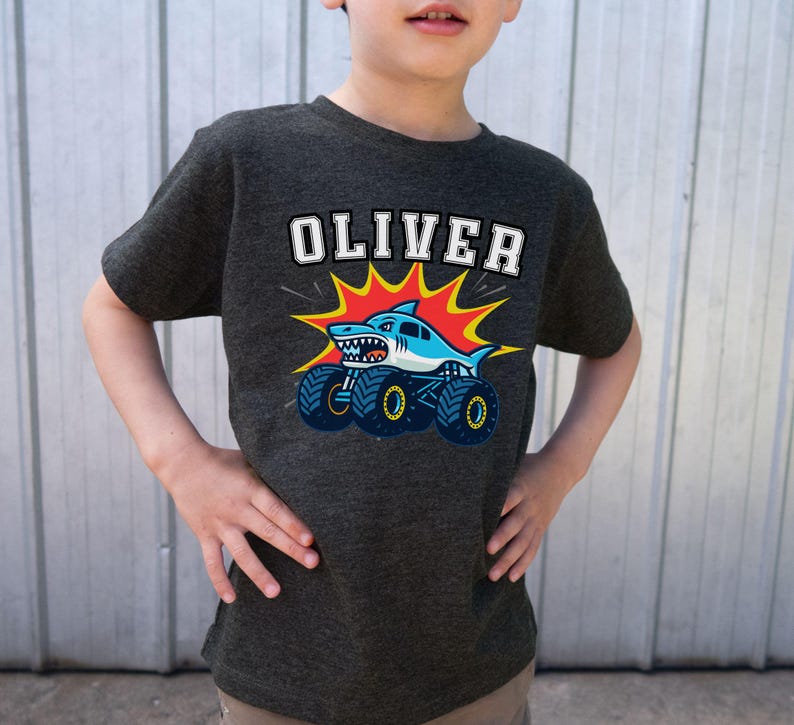 May include: Dark gray t-shirt featuring the name "OLIVER" in white block letters above a cartoon shark monster truck design. The shark is blue and white with sharp teeth, and the truck has large blue wheels. The design is set against a red and yellow burst.