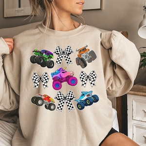 Puede incluir: Sudadera beige con gráficos de monster trucks coloridos y lazos a cuadros. Los camiones son de varios colores, incluyendo verde, naranja, rojo y azul, con un camión unicornio en el centro. El diseño es lúdico y llamativo.
