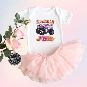 Puede incluir: Un body blanco para beb&eacute; con un gr&aacute;fico de monster truck colorido y las palabras "Kidster Jam". Combinado con una falda tut&uacute; rosa claro. El dise&ntilde;o incluye una bandera a cuadros y una fuente l&uacute;dica.