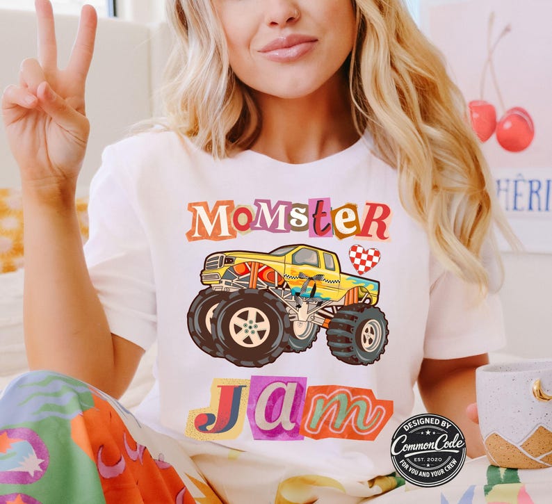 Pictured is a woman wearing a natural off white t-shirt which says Momster Jam in scrapbook letters along with a yellow multi-colored monster truck with a red and white checkered heart
