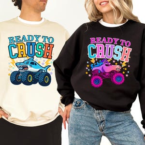 Pu&ograve; includere: Felpe beige e nere con la scritta "READY TO CRUSH". La felpa beige presenta un design di monster truck a forma di squalo, mentre la felpa nera ha un design di monster truck a forma di unicorno. Entrambe le felpe hanno grafiche colorate.