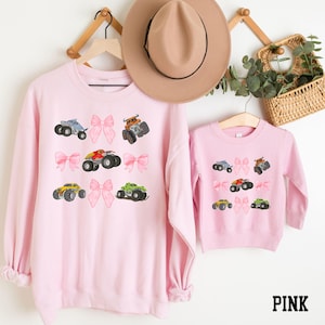 May include: Pink sweatshirts with monster truck and bow designs. The adult sweatshirt has a crew neck and long sleeves. The children's sweatshirt has a crew neck and long sleeves. The word "PINK" is printed in black.