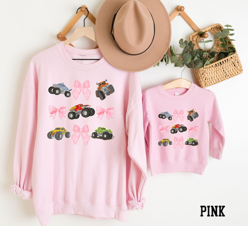 May include: Pink sweatshirts with monster truck and bow designs. The adult sweatshirt has a crew neck and long sleeves. The children's sweatshirt has a crew neck and long sleeves. The word "PINK" is printed in black.