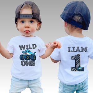 Op de afbeelding: Een wit T-shirt met een haai-thema monstertruck-afbeelding en de tekst "WILD ONE". De achterkant heeft de naam "LIAM" en het nummer "1" met een monstertruck-afbeelding. Het kind draagt een pet.