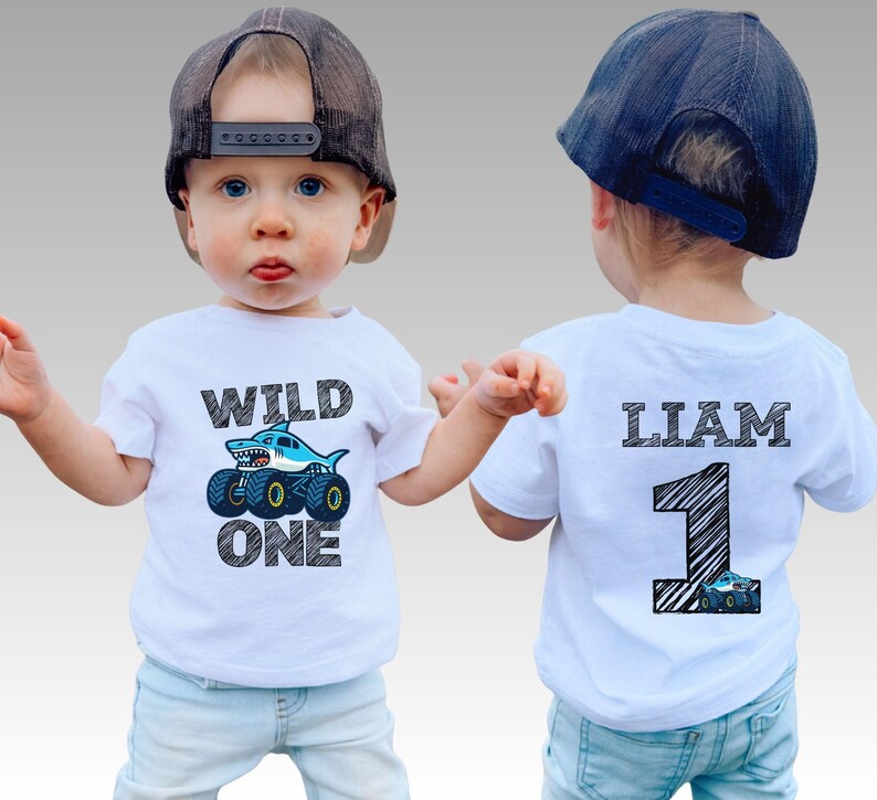 Op de afbeelding: Een wit T-shirt met een haai-thema monstertruck-afbeelding en de tekst "WILD ONE". De achterkant heeft de naam "LIAM" en het nummer "1" met een monstertruck-afbeelding. Het kind draagt een pet.