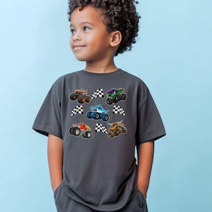 May include: A charcoal gray t-shirt featuring colorful monster truck graphics and checkered flags. The trucks are in various colors, including orange, green, blue, and red. The shirt is worn by a young child.