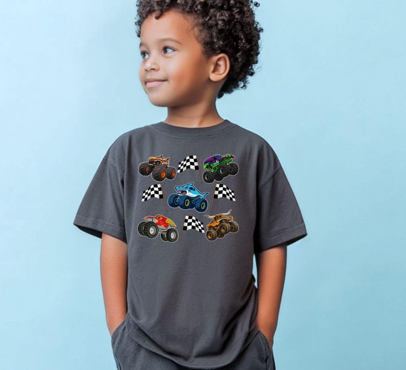 May include: A charcoal gray t-shirt featuring colorful monster truck graphics and checkered flags. The trucks are in various colors, including orange, green, blue, and red. The shirt is worn by a young child.