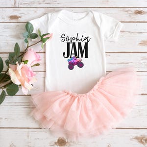 May include: A white baby bodysuit with the text "Sophia JAM" and a colorful monster truck graphic. A light pink tutu skirt is paired with the bodysuit. The outfit is designed for a baby or toddler.