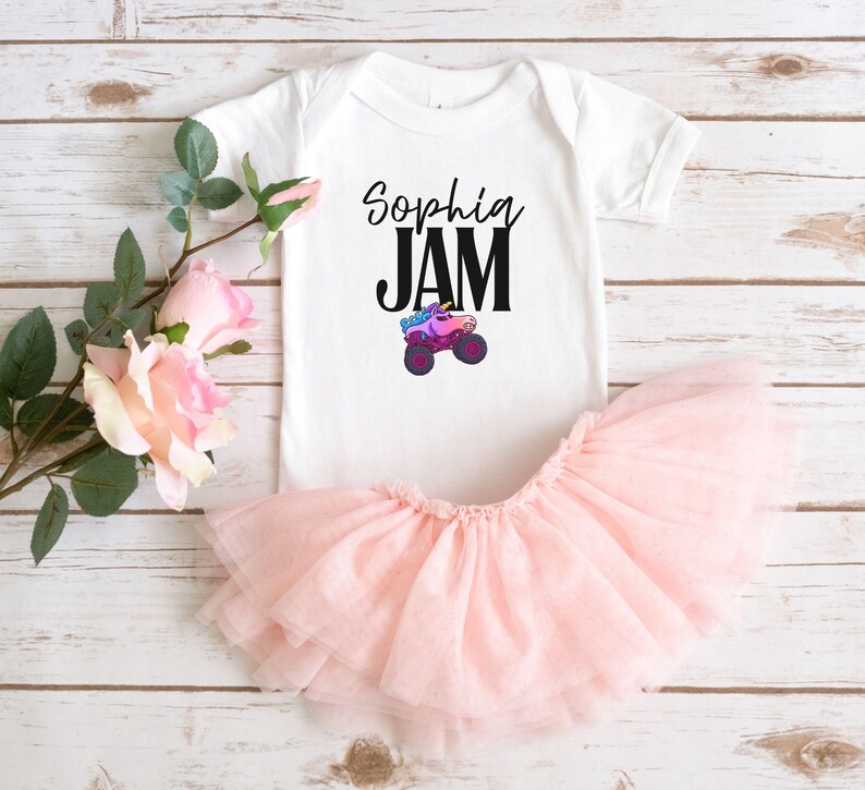 May include: A white baby bodysuit with the text "Sophia JAM" and a colorful monster truck graphic. A light pink tutu skirt is paired with the bodysuit. The outfit is designed for a baby or toddler.