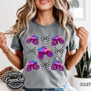 May include: Grey t-shirt with a pattern of monster trucks with unicorn horns and checkered bows. The trucks are pink, purple, and blue with large wheels. The text "Designed by Common Code Est. 2020 For You and Your Crew" is visible.