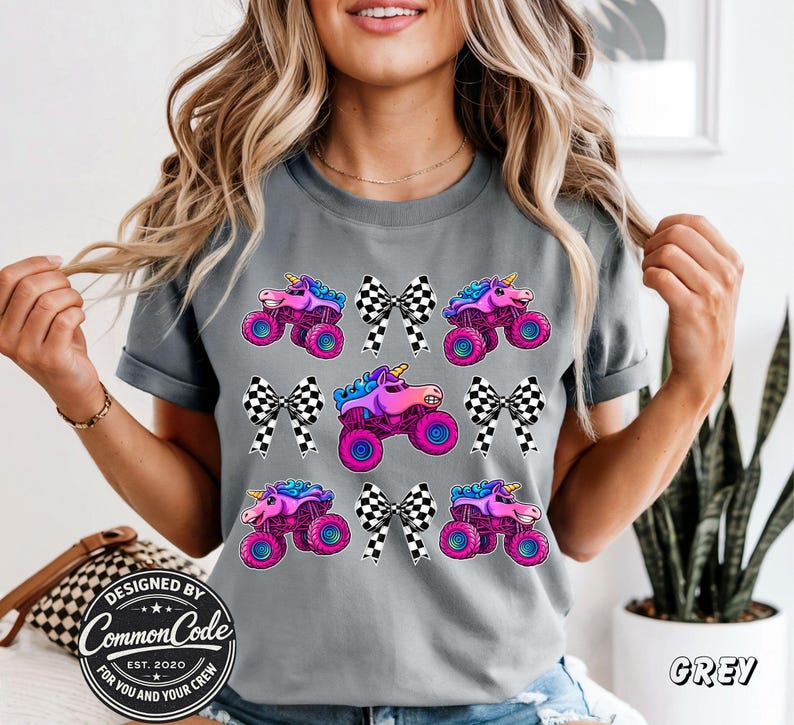 May include: Grey t-shirt with a pattern of monster trucks with unicorn horns and checkered bows. The trucks are pink, purple, and blue with large wheels. The text "Designed by Common Code Est. 2020 For You and Your Crew" is visible.