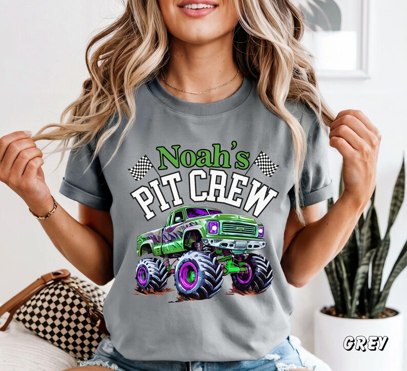 Pode incluir: Camiseta cinza com um gr&aacute;fico de um monster truck verde com rodas roxas e pretas. O texto "Noah's PIT CREW" est&aacute; acima do caminh&atilde;o em verde e branco. Bandeiras quadriculadas est&atilde;o em ambos os lados do texto.