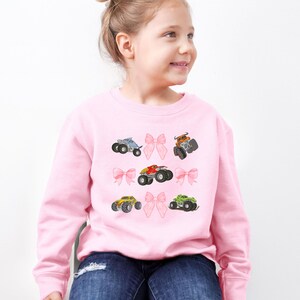 May include: A light pink sweatshirt featuring colorful monster truck graphics and pink bow accents. The design includes trucks in blue, orange, red, yellow, and green. A fun and playful design for kids.