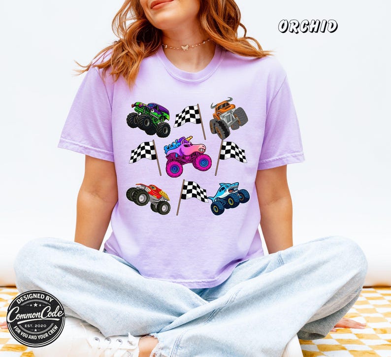 May include: A light purple t-shirt with a graphic of colorful monster trucks and checkered flags. The shirt has the word "ORCHID" printed at the top. The bottom left corner of the shirt has the text "DESIGNED BY COMMON CODE EST. 2020 FOR YOU AND YOUR CREW".