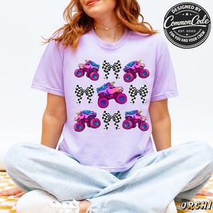May include: A lilac t-shirt with a repeating pattern of unicorn monster trucks and black and white checkered bows. The shirt has the text "DESIGNED BY Common Code EST. 2020 FOR YOU AND YOUR CREW" in a circular design.