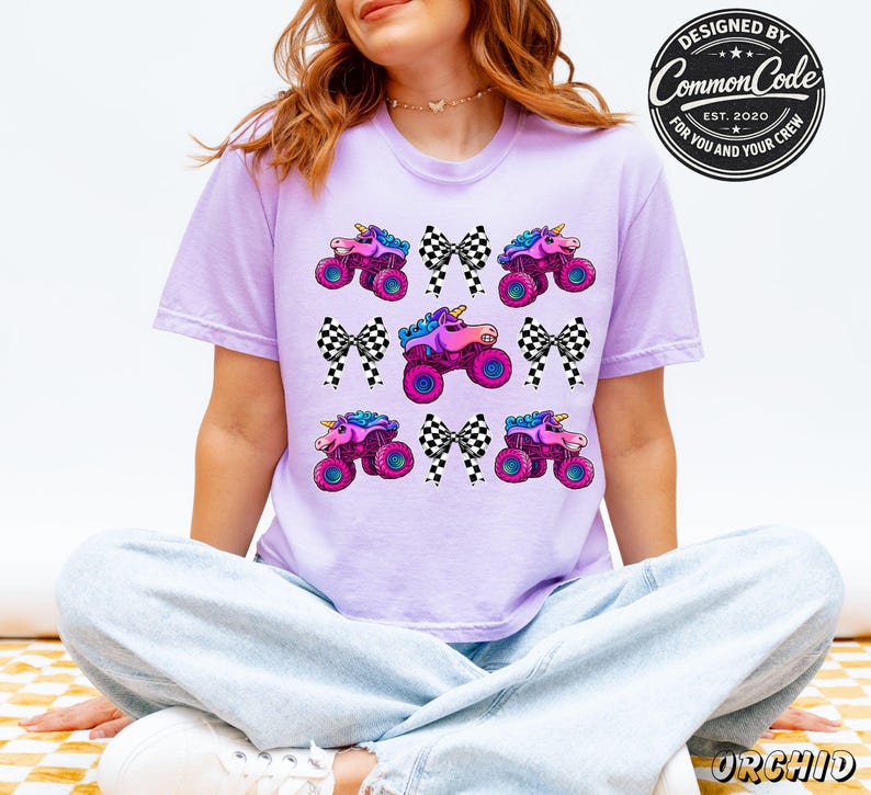 May include: A lilac t-shirt with a repeating pattern of unicorn monster trucks and black and white checkered bows. The shirt has the text "DESIGNED BY Common Code EST. 2020 FOR YOU AND YOUR CREW" in a circular design.