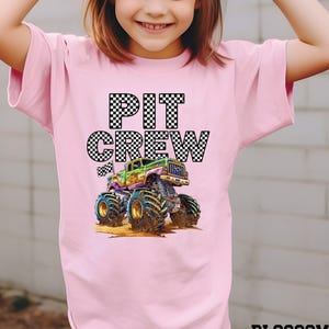 May include: Pink t-shirt featuring the words "PIT CREW" in a checkered pattern above a colorful monster truck graphic. The monster truck is green, purple, and yellow with large tires. The word "BLOSSOM" is at the bottom right.