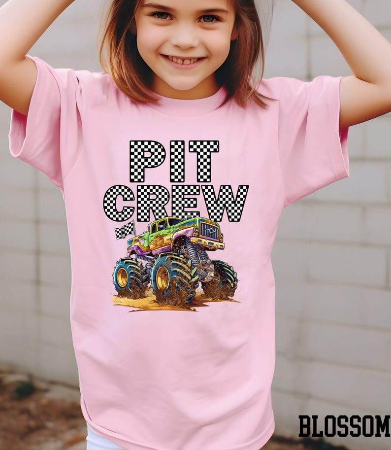 May include: Pink t-shirt featuring the words "PIT CREW" in a checkered pattern above a colorful monster truck graphic. The monster truck is green, purple, and yellow with large tires. The word "BLOSSOM" is at the bottom right.