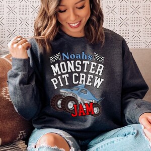 May include: Dark gray sweatshirt with the text "Noah's MONSTER PIT CREW JAM" in white, blue, and red. The design features a cartoon shark monster truck with large tires and checkered flags.