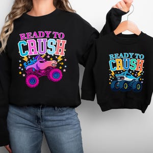 Mommy and Mini Me Monster Truck Racing Theme sweatshirts. The mama is wearing a black sweatshirt holding a black toddler sweatshirt that matches. Hers says" READY TO CRUSH" in pinks, purples and blues with a sparkle unicorn smash truck with stars and lightning. The toddler sweatshirt says "READY TO CRUSH" Varsity style in blues and oranges with a shark megalodon monster truck with stars and lightning. Perfect Boy Mama and Mama's boy as well as Girl Mama - choose your design on each sweater.