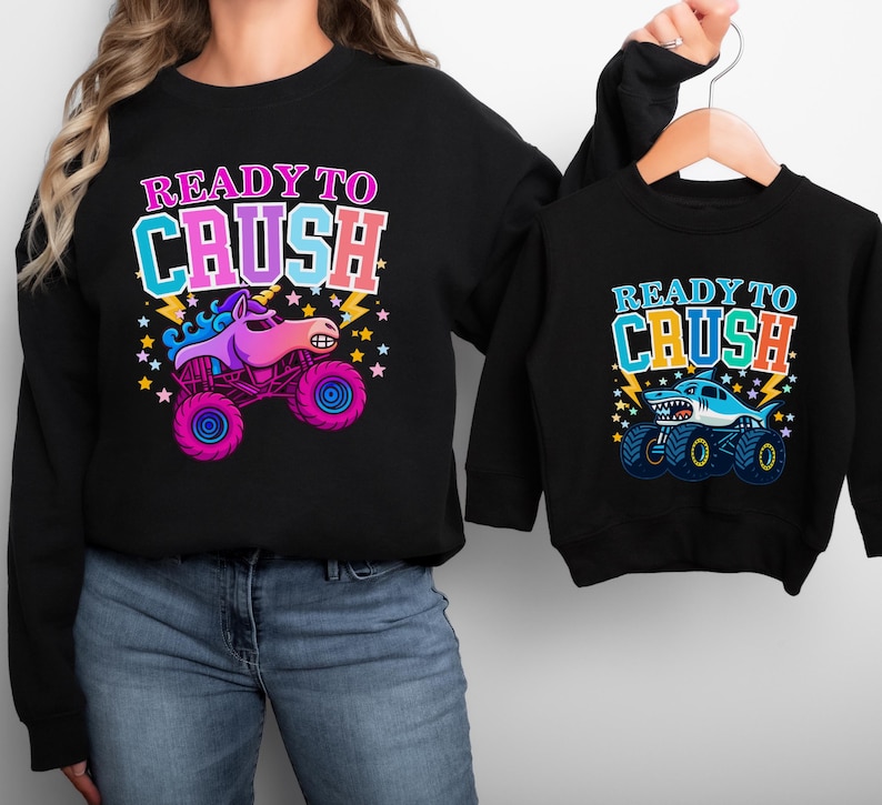 Mommy and Mini Me Monster Truck Racing Theme sweatshirts. The mama is wearing a black sweatshirt holding a black toddler sweatshirt that matches. Hers says" READY TO CRUSH" in pinks, purples and blues with a sparkle unicorn smash truck with stars and lightning. The toddler sweatshirt says "READY TO CRUSH" Varsity style in blues and oranges with a shark megalodon monster truck with stars and lightning. Perfect Boy Mama and Mama's boy as well as Girl Mama - choose your design on each sweater.
