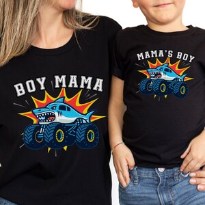May include: Black t-shirts with matching graphics. One shirt reads "BOY MAMA" with a shark monster truck design. The other shirt says "MAMA'S BOY" with a similar shark monster truck design. Both shirts have a blue, white, and yellow color scheme.