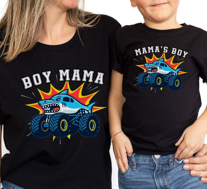 May include: Black t-shirts with matching graphics. One shirt reads "BOY MAMA" with a shark monster truck design. The other shirt says "MAMA'S BOY" with a similar shark monster truck design. Both shirts have a blue, white, and yellow color scheme.